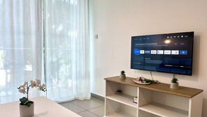 A modern living room with a flat-screen TV mounted on the wall, a white table with a potted orchid, and a wooden shelf unit. - 122 PRO - Stay Tropical Apartment (Paralimni)