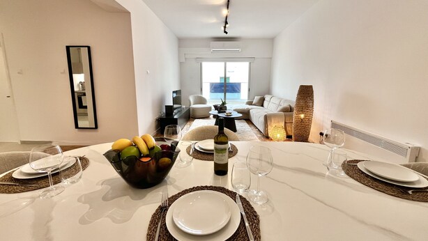 Un comedor con una mesa preparada para dos personas, un cuenco de fruta y una botella de vino.