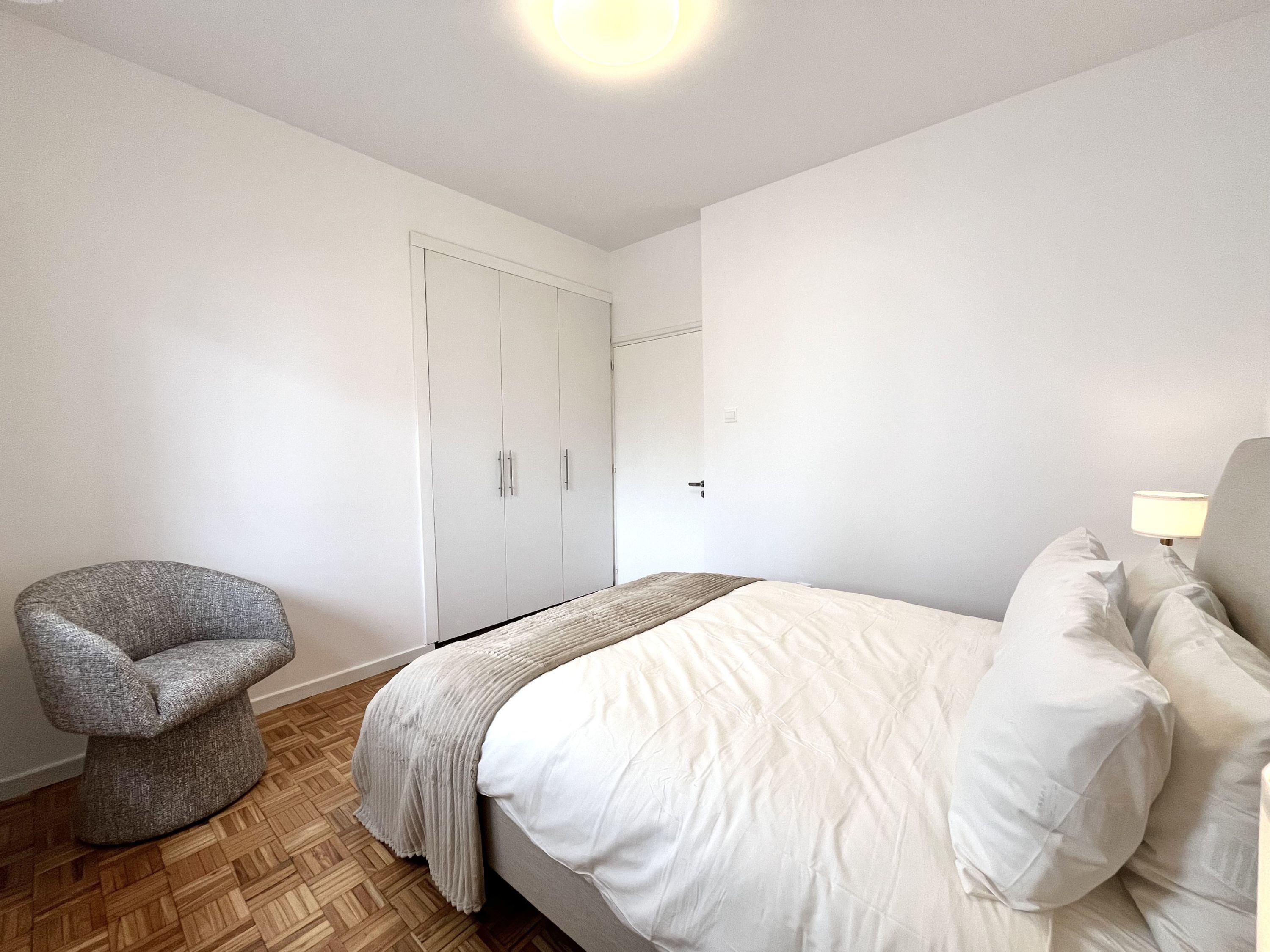 Un dormitorio con una cama, una silla, un armario y una mesita de noche con lámpara.