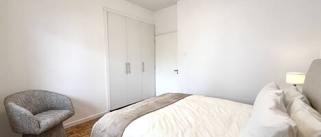 Un dormitorio con una cama, una silla, un armario y una mesita de noche con lámpara.