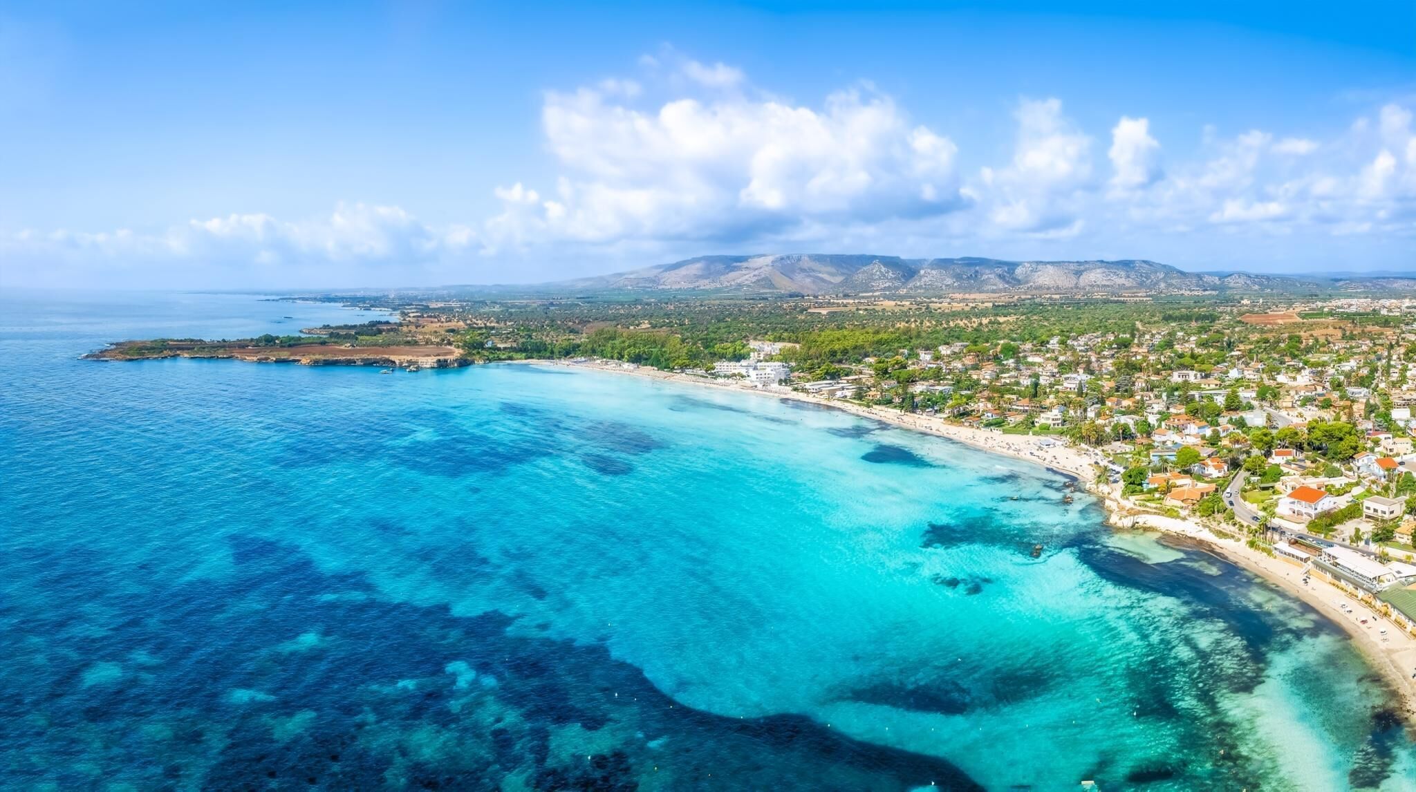 An aerial view of a coastal town with clear blue waters, a beach, and a mountainous backdrop.