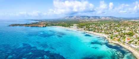 Une vue aérienne d’une ville côtière, avec des eaux bleues et limpides, une plage et un paysage montagneux en arrière-plan.