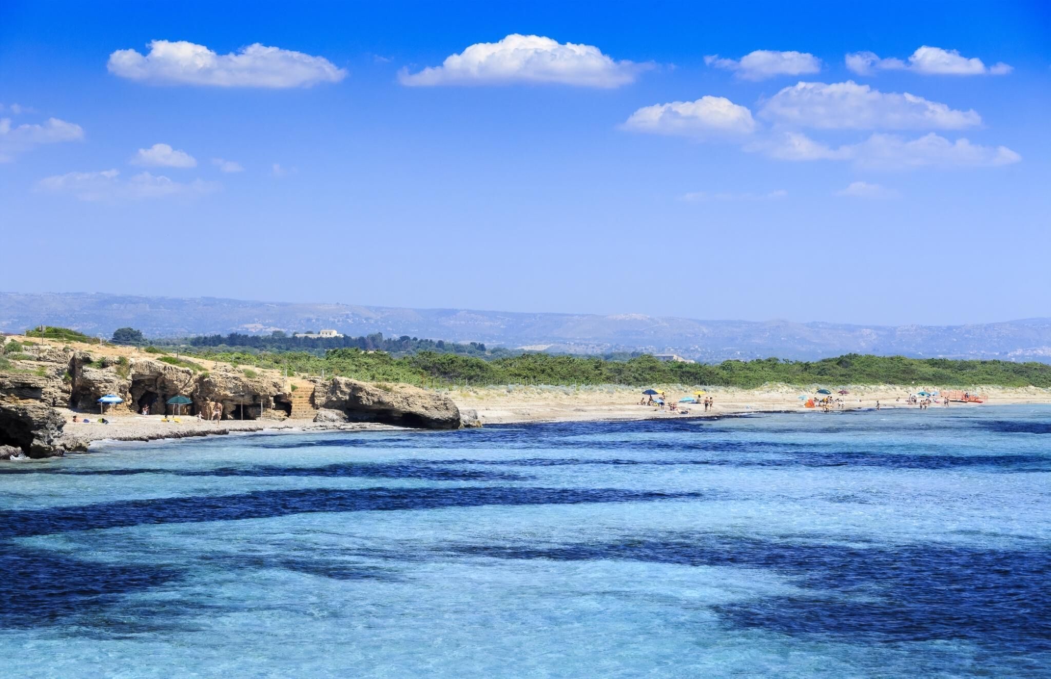 Un paysage côtier caractérisé par une eau d’un bleu limpide, des affleurements rocheux et une vue lointaine sur des collines.