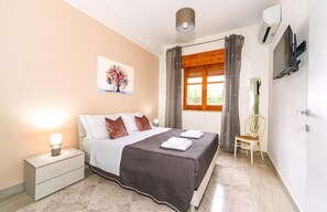 Un dormitorio con cama, mesita de noche, lámpara, obra de arte, ventana con cortinas y una silla.