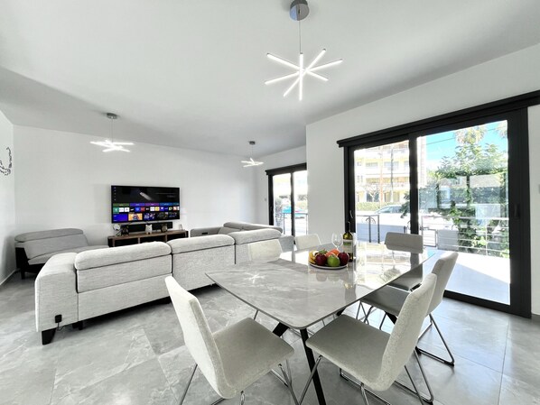 Ein modernes Wohnzimmer mit Flachbildfernseher, grauem Sofa, Esstisch mit Stühlen und großen Fenstern.