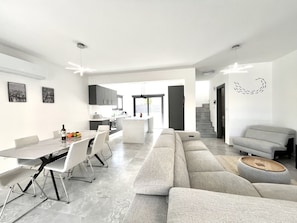A modern kitchen with dark cabinets, a dining area with a long table and chairs, a grey sofa, and a staircase leading to another level.