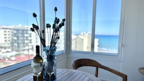 A round wooden table with a wine bottle and glasses, a vase with dried flowers, and a view of the ocean and buildings through large windows. - 39 LCA - Stay Makenzie View Apartment (Larnaca)