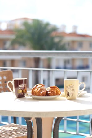 A table with two coffee mugs and a plate of croissants on a balcony with a railing and a building in the background. - 43 PAP - Stay Iris Apartment (Paphos)
