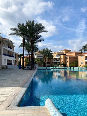 A swimming pool with clear blue water, surrounded by palm trees and multi-story buildings with balconies. - 43 PAP - Stay Iris Apartment (Paphos)