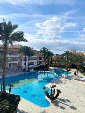A swimming pool with a curved design, surrounded by palm trees and a building complex with balconies. - 43 PAP - Stay Iris Apartment (Paphos)