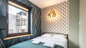 Una cama con un edredón verde, dos almohadas blancas y una ventana con vistas a edificios.