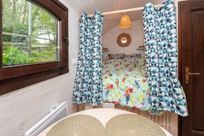 Un dormitorio con piso de madera, una cama con colcha estampada, una ventana con cortinas, una puerta y una lámpara de pared.
