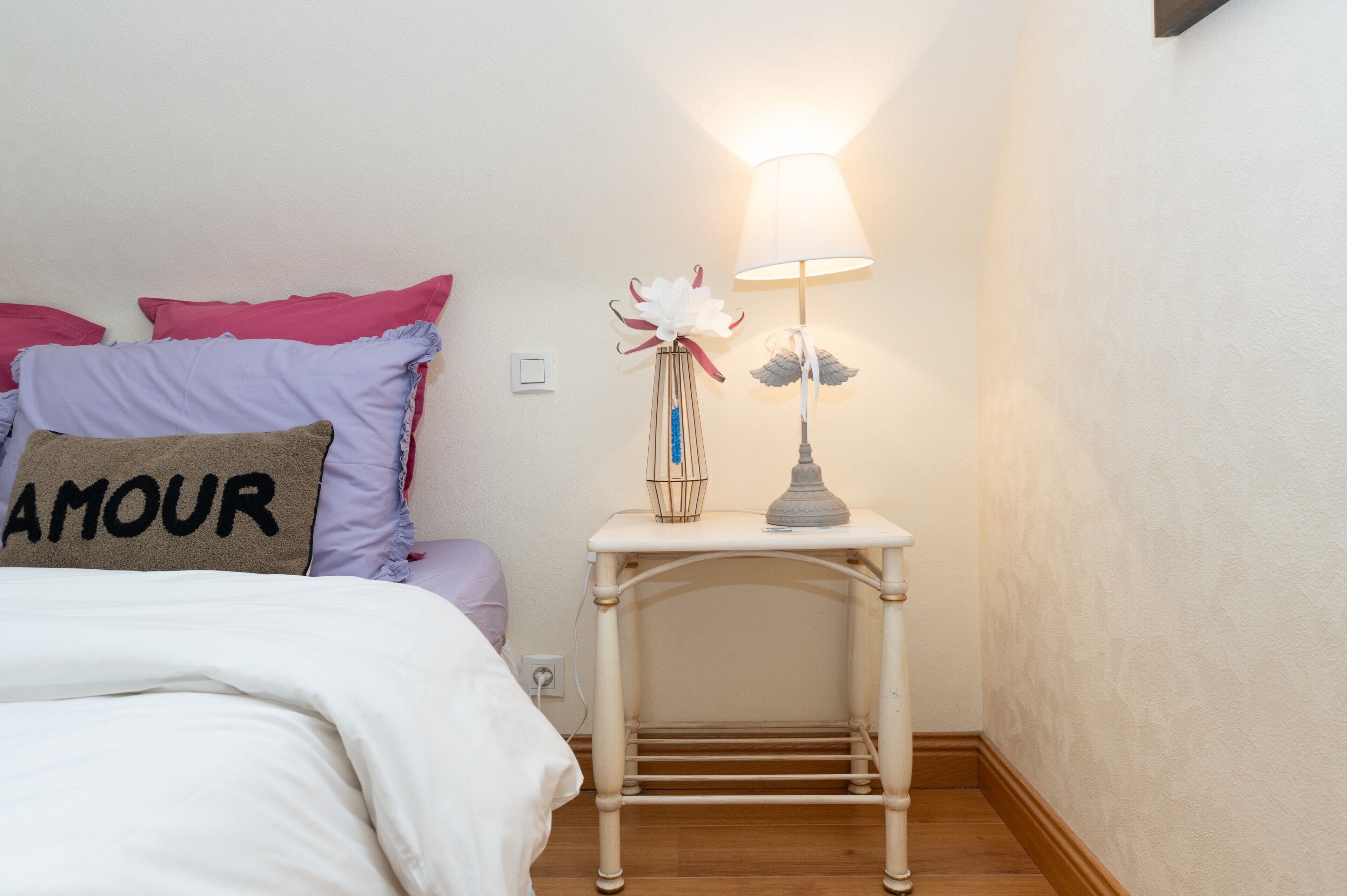 A neatly made bed with a pillow labeled 'AMOUR,' a bedside table with a lamp, and a wall switch.