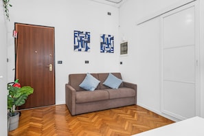 A room with a wooden door, a brown sofa with two pillows, a potted plant, and a herringbone-patterned floor. - "3 Beds Duomo Square "Modern House+6 Pax By KeyHost (Milano)