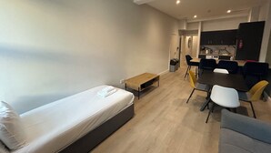 Une chambre moderne avec un lit, une petite table, un coin repas avec des chaises et, au fond, une cuisine.