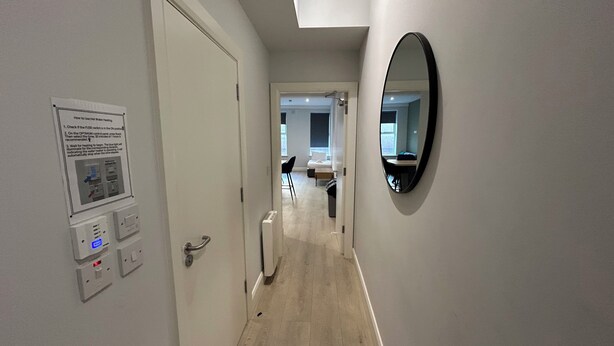 Un couloir avec une porte, un panneau de commande fixé au mur, un radiateur et un miroir.