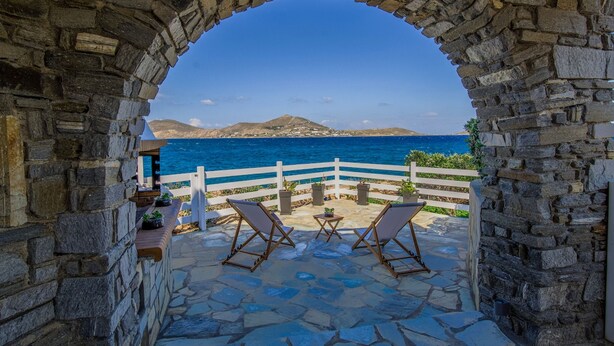 Un arco de piedra que da a una terraza con dos sillones y una mesita, con vistas a la costa, una valla blanca y una montaña distante.