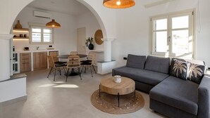 A modern living room with a grey sofa, a round wooden coffee table, and a dining area with wicker chairs. - Haven in Naousa (Paros)