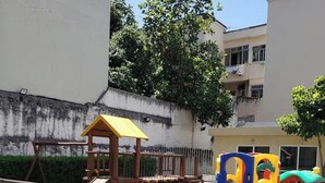 Une aire de jeux avec une structure d’escalade jaune et un toboggan coloré, entourée d’une clôture et d’une zone dallée.