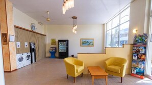 A hotel lobby with a reception desk, two yellow armchairs, a small table, a washing machine, a refrigerator, and a large window. - Only Room Hotel  (Lido di Savio)