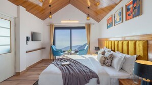 Habitación de hotel con una cama grande, televisión, ventana con vistas a la montaña y zona de comedor con sillas.