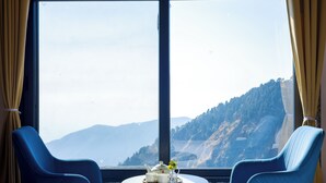 Una habitación con dos sillas azules, una pequeña mesa redonda con una tetera y tazas, y un gran ventanal con vistas a la montaña.