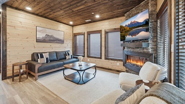 A cozy living room with a stone fireplace, wooden walls, and a large window with blinds. - NEW Build - Smokies Haven w/ Hot Tub, Games, MORE (Pigeon Forge)