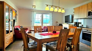 A modern kitchen with a dining area, wooden cabinets, a TV, and a large window.