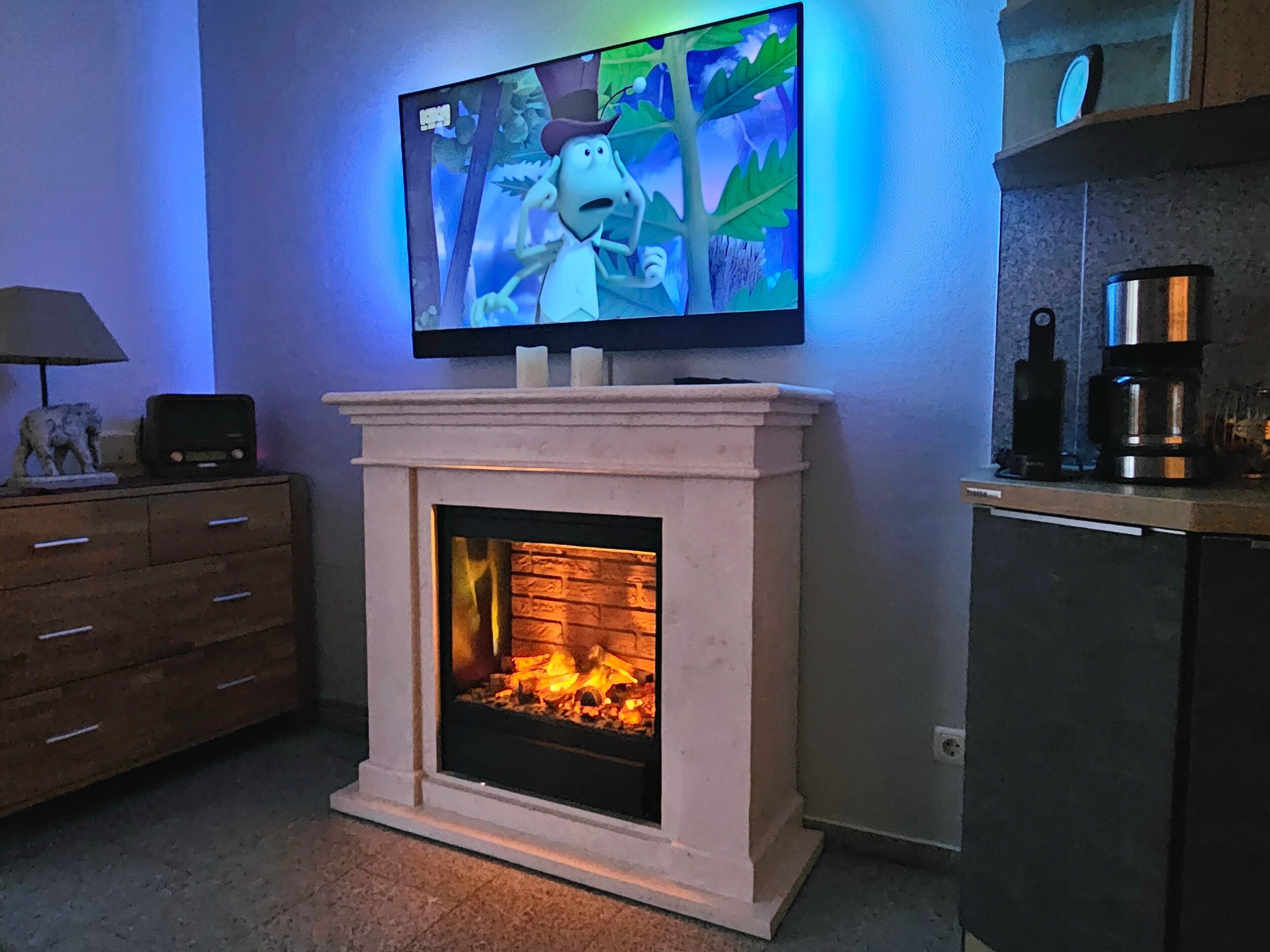 A fireplace with a TV mounted above it, a wooden dresser to the left, and a coffee machine to the right.