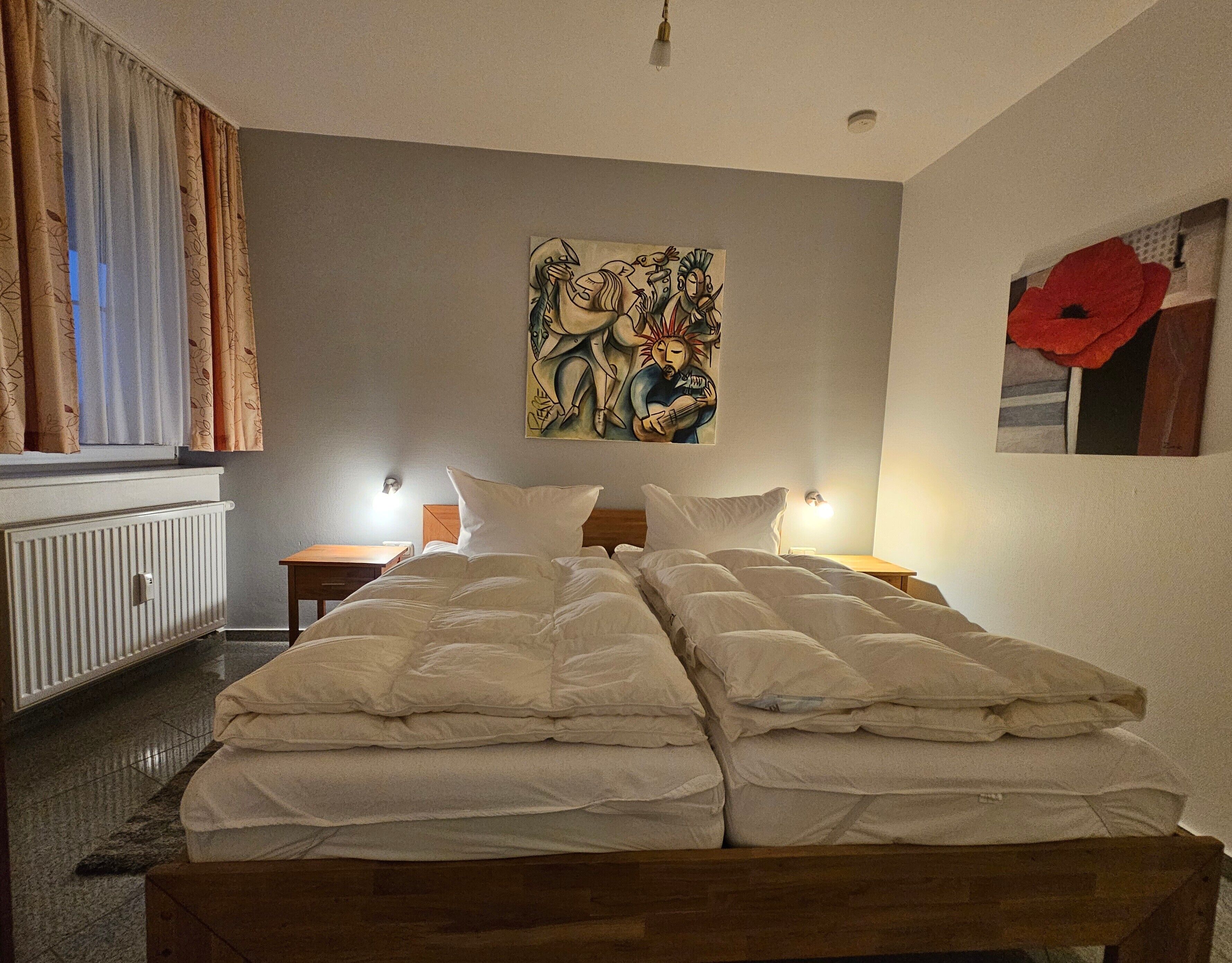 Ein Hotelzimmer mit Doppelbett, zwei Nachttischen mit Lampen, einem Heizkörper, einem Fenster mit Vorhängen und einem Bild an der Wand.