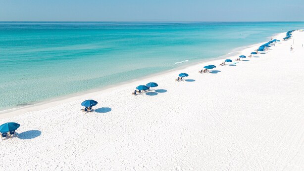Een strand met wit zand en helder turquoise water. Er staan rijen blauwe parasols en strandstoelen.