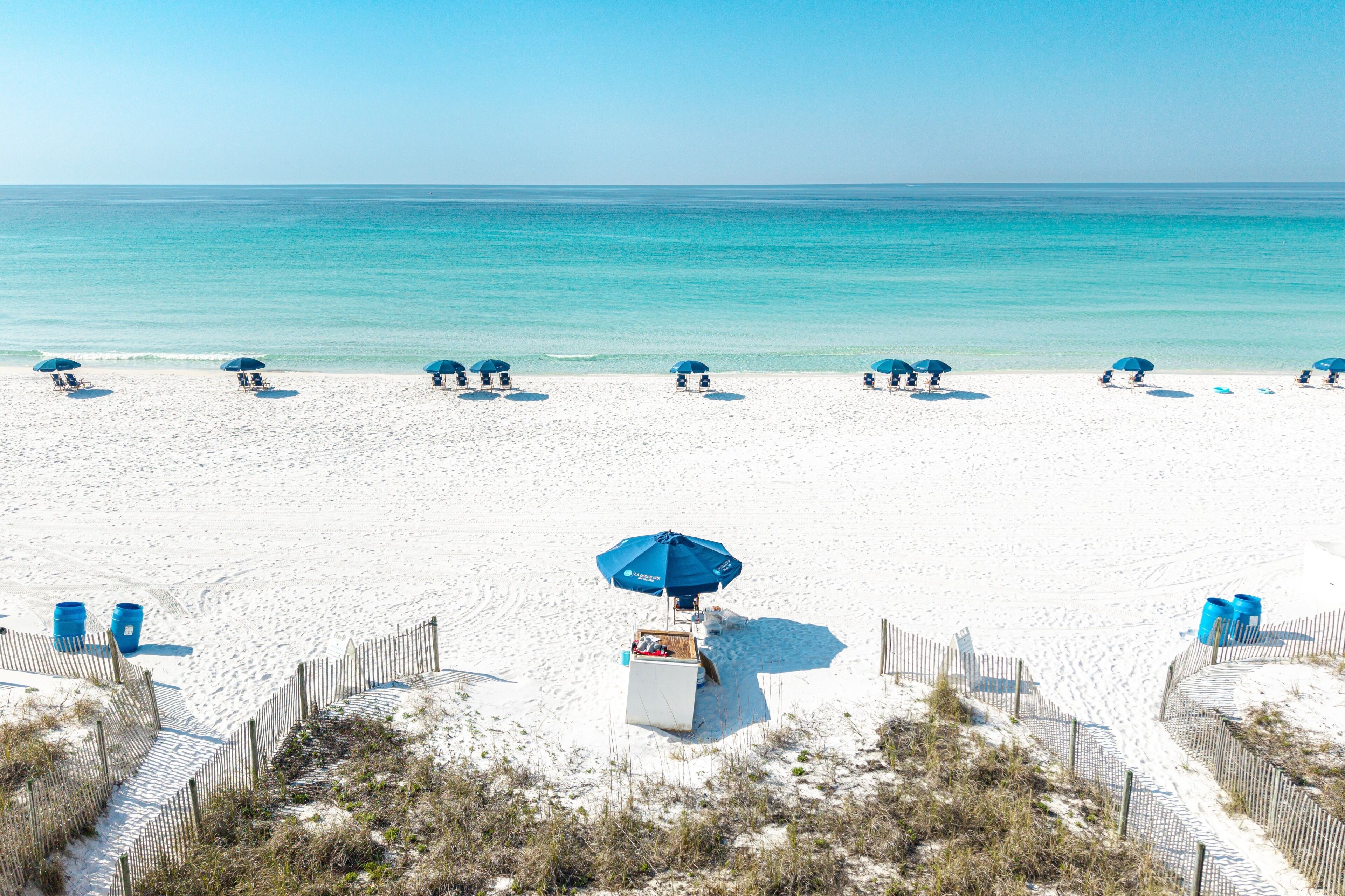 A beach with white sand, a wooden fence, and a row of blue umbrellas.