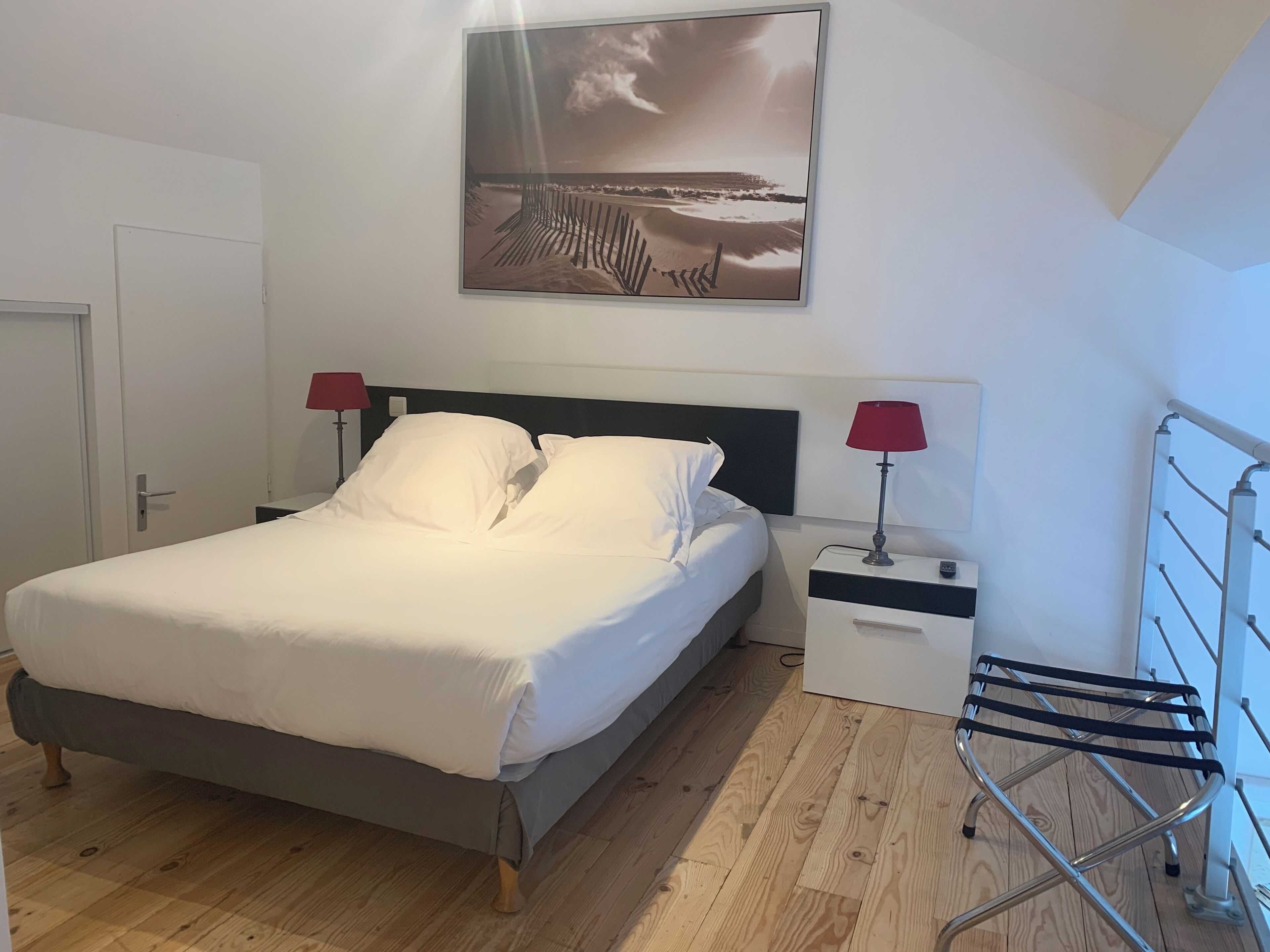 A bedroom with a bed, two bedside tables with lamps, a wooden floor, a door, and a wall-mounted artwork.