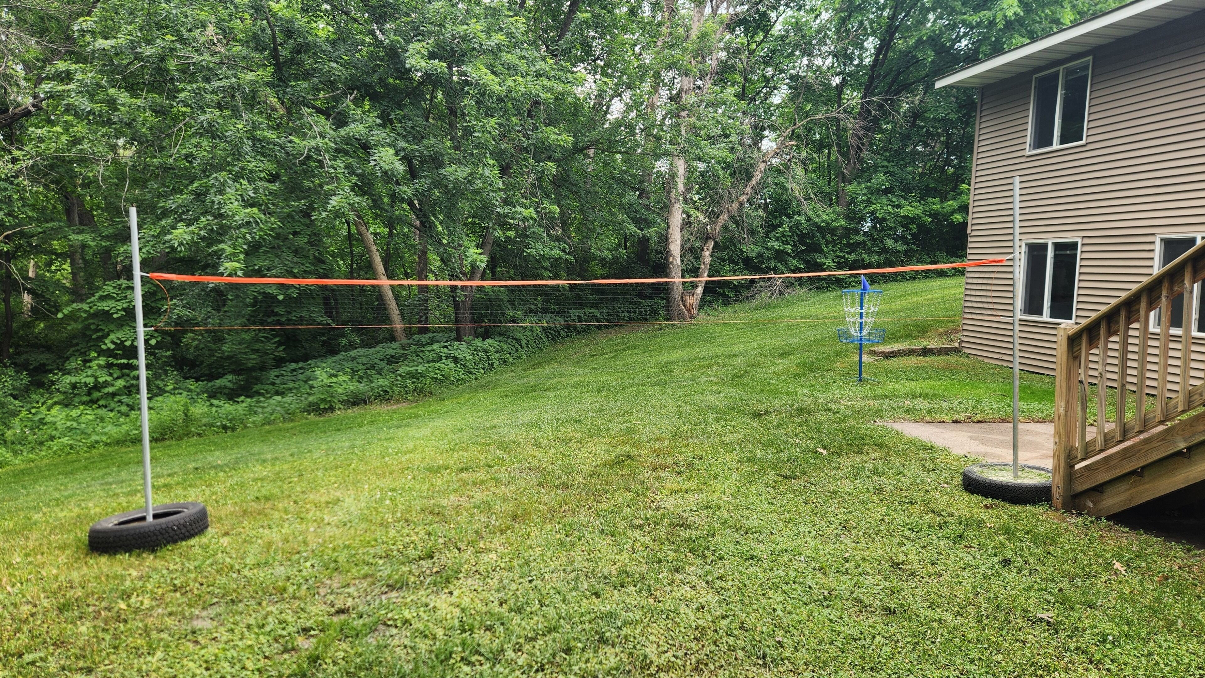 Une cour arrière verdoyante avec une balançoire faite d’un pneu, un panier bleu pour le disc golf et une terrasse en bois.