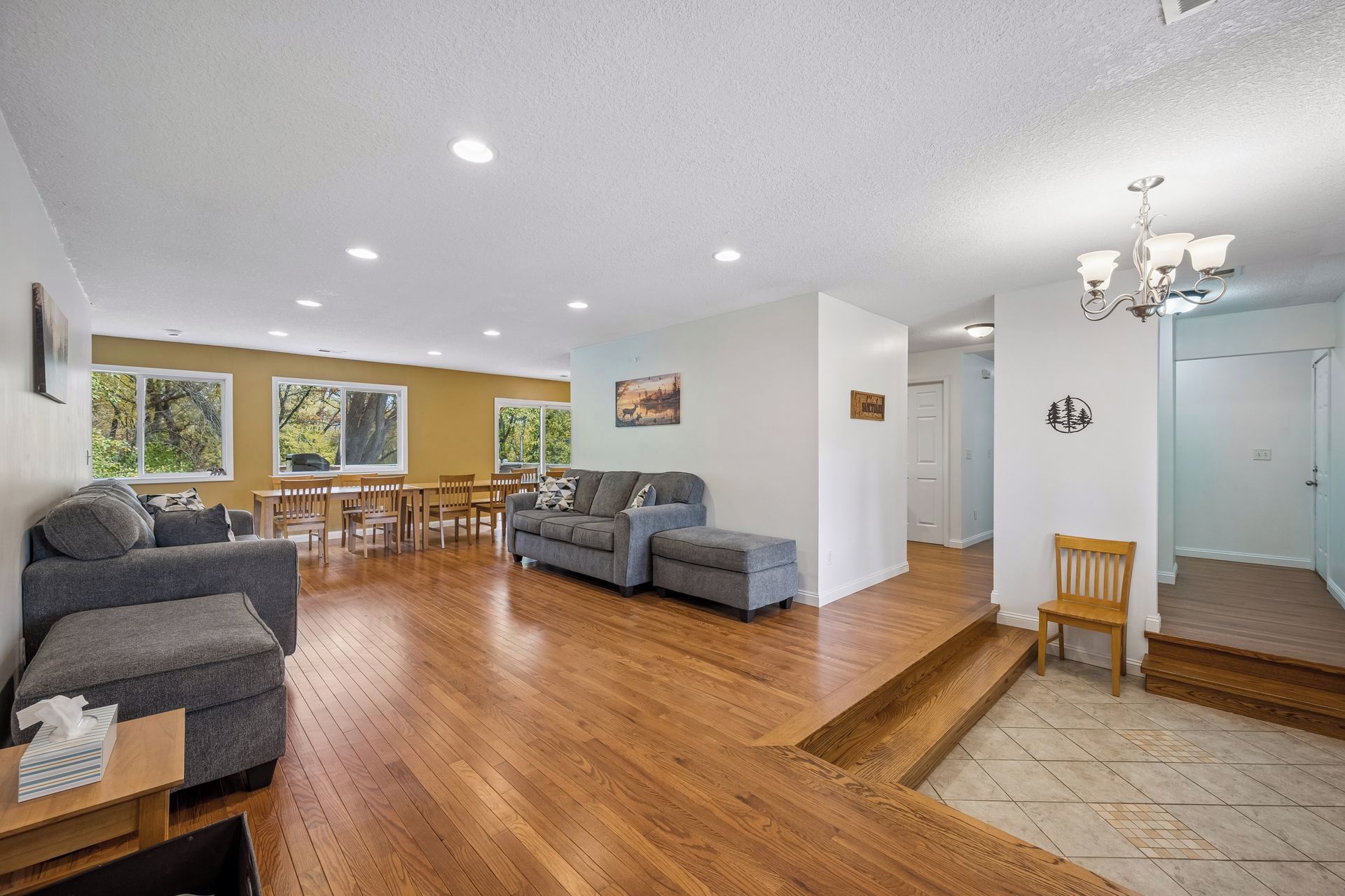 A spacious living area with a wooden floor, a gray sofa, a dining table with chairs, and a staircase leading to another level.