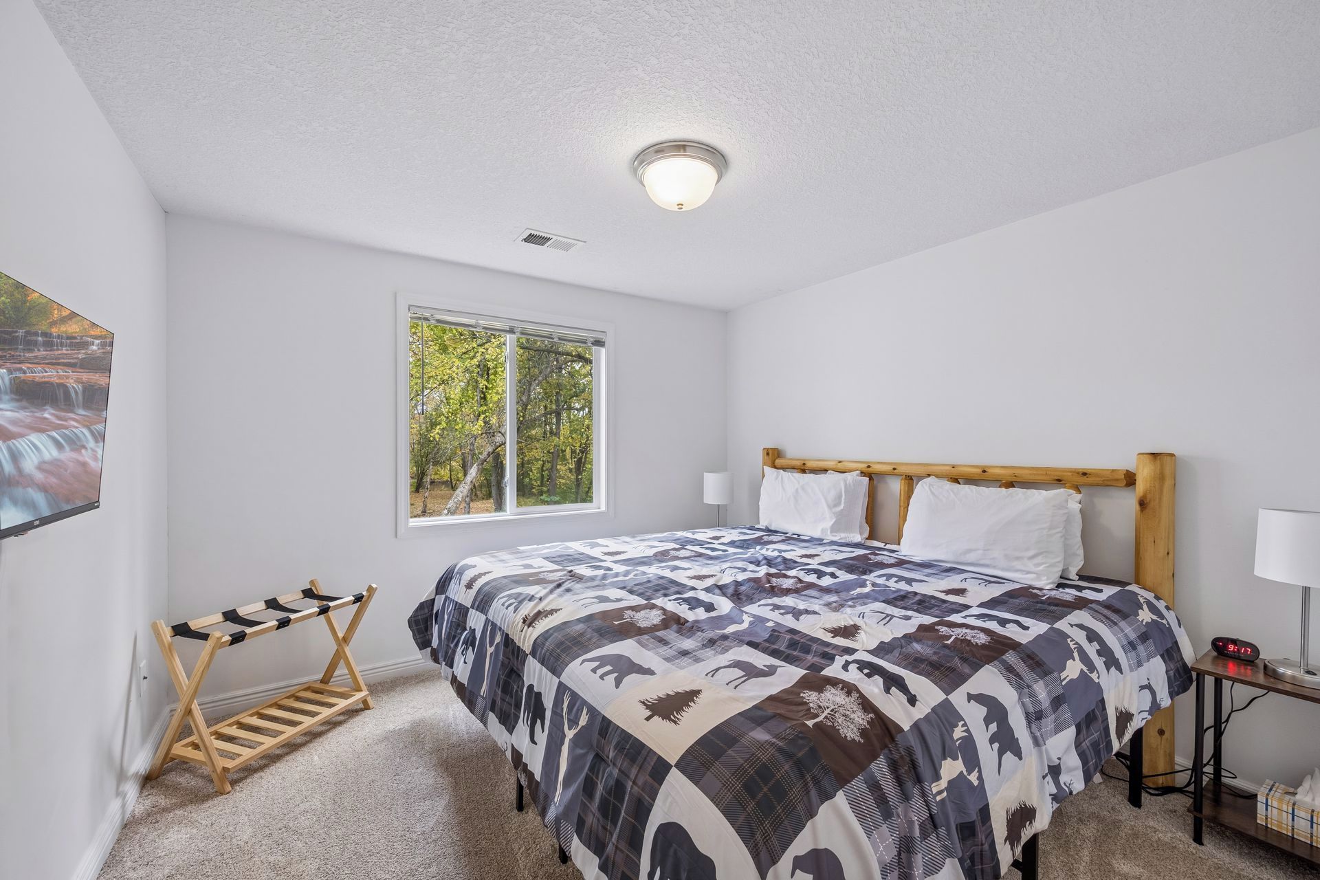 A bedroom with a bed, a window, a ceiling light, a wall-mounted TV, and a bedside table with a lamp.