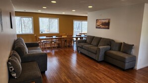Un salon spacieux avec un canapé gris, un coin repas avec des chaises en bois et une table, et de grandes fenêtres donnant sur la forêt.