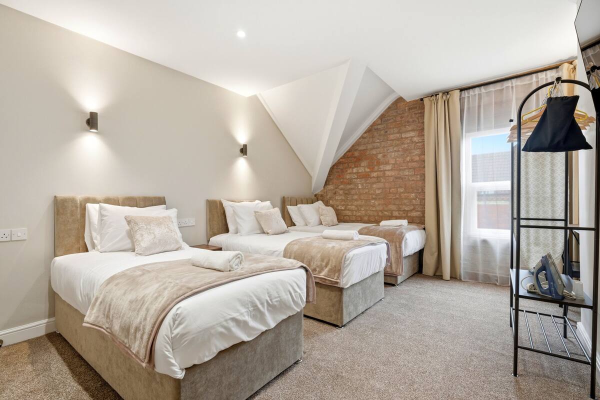A hotel room with three beds, a brick wall, a window with curtains, and a metal rack with items.