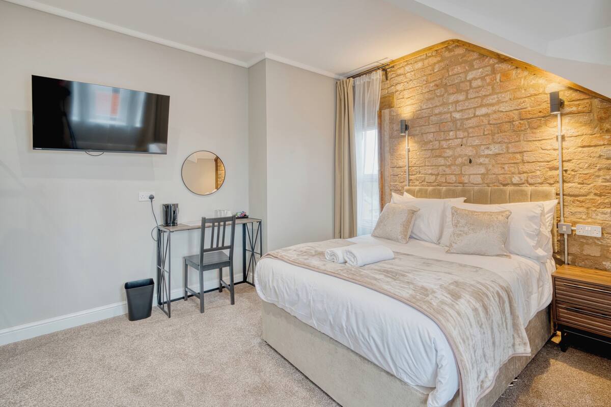 A bedroom with a large bed, a desk, a chair, a TV, and a stone wall.