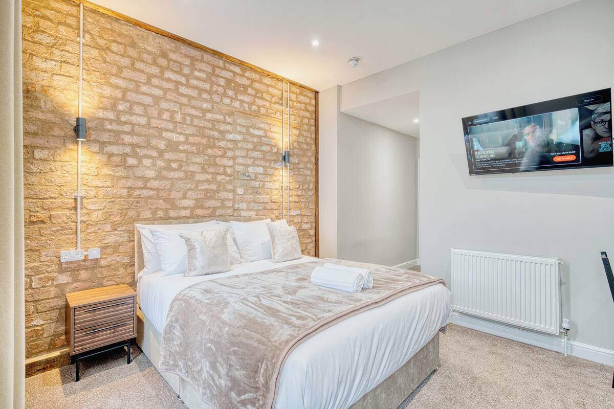 A modern bedroom with a brick accent wall, a bed with white linens, a mounted TV, and a white radiator.