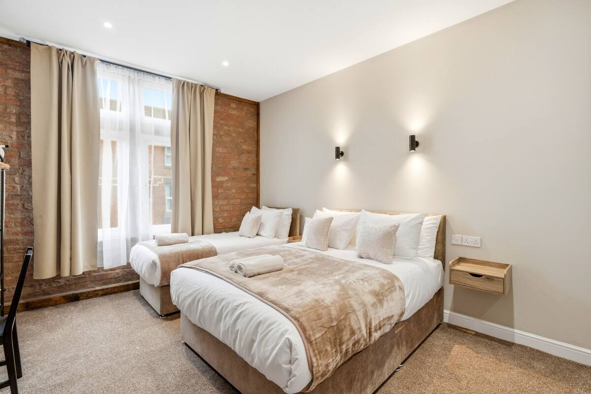 A hotel room with two beds, a brick wall, a window with curtains, and a nightstand with a drawer.