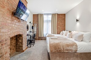 A hotel room with a brick fireplace, a flat-screen TV mounted on the wall, a bed with white linens, a table with chairs, and a window with curtains. - The Stuart Hotel (Liverpool)