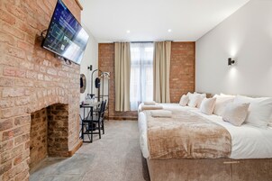 Habitación de hotel con chimenea de ladrillo, un televisor de pantalla plana montado en la pared, una cama con ropa blanca, una mesa con sillas y una ventana con cortinas.