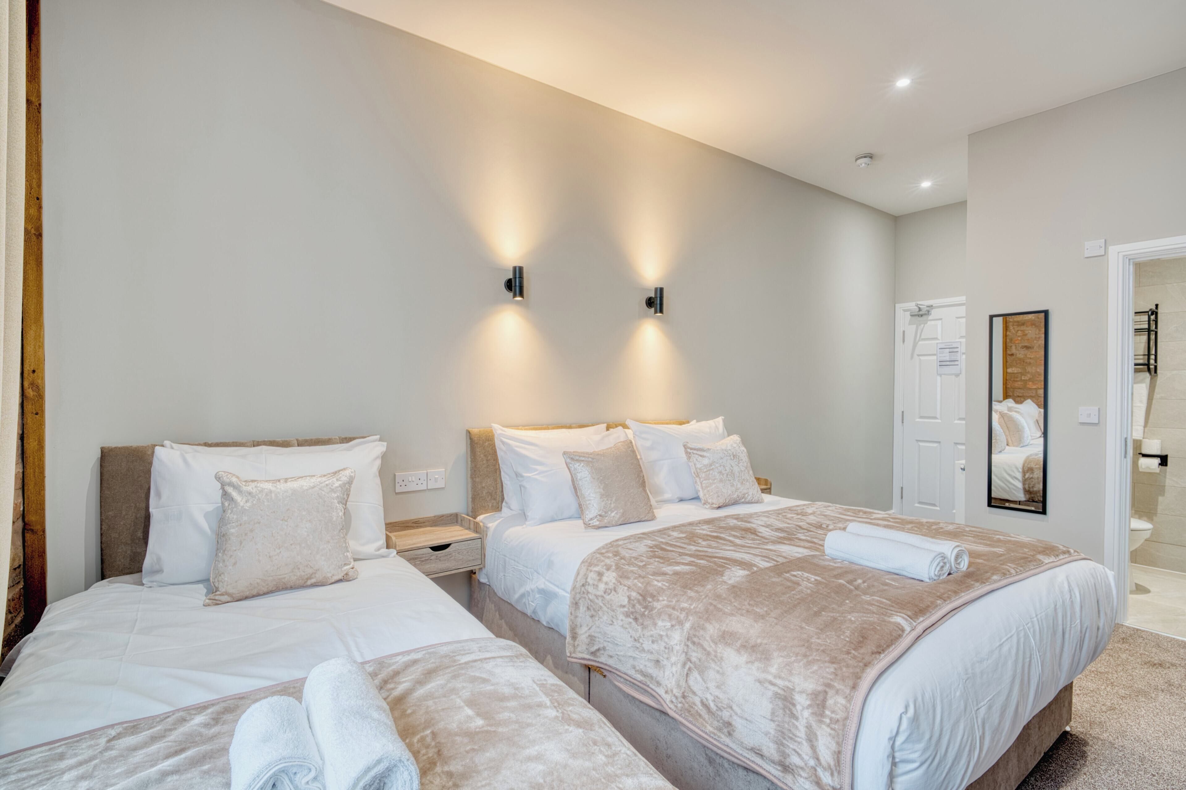 A hotel room with two beds, a nightstand, a wall-mounted light fixture, a door, and a mirror.