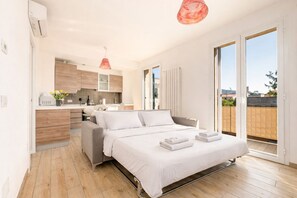 Un dormitorio moderno con cama, una pequeña zona de estar, una cocina al fondo y un ventanal corredero al exterior.