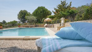 A swimming pool with a wicker lounge chair and a cushion, surrounded by pebbles and trees. - Les Olivandes Cottage with Pool (Saint-Cannat)