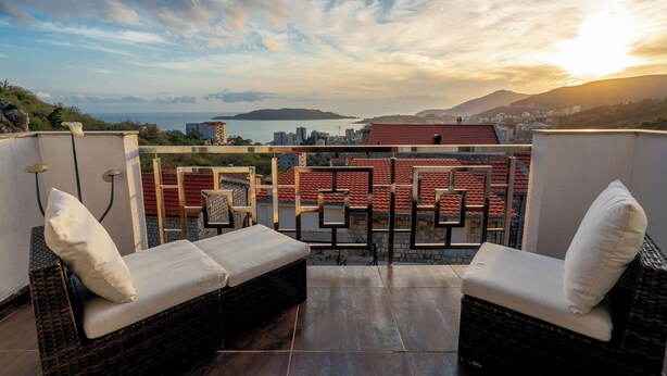 A balcony with wicker furniture, a view of the sea, and buildings in the background.