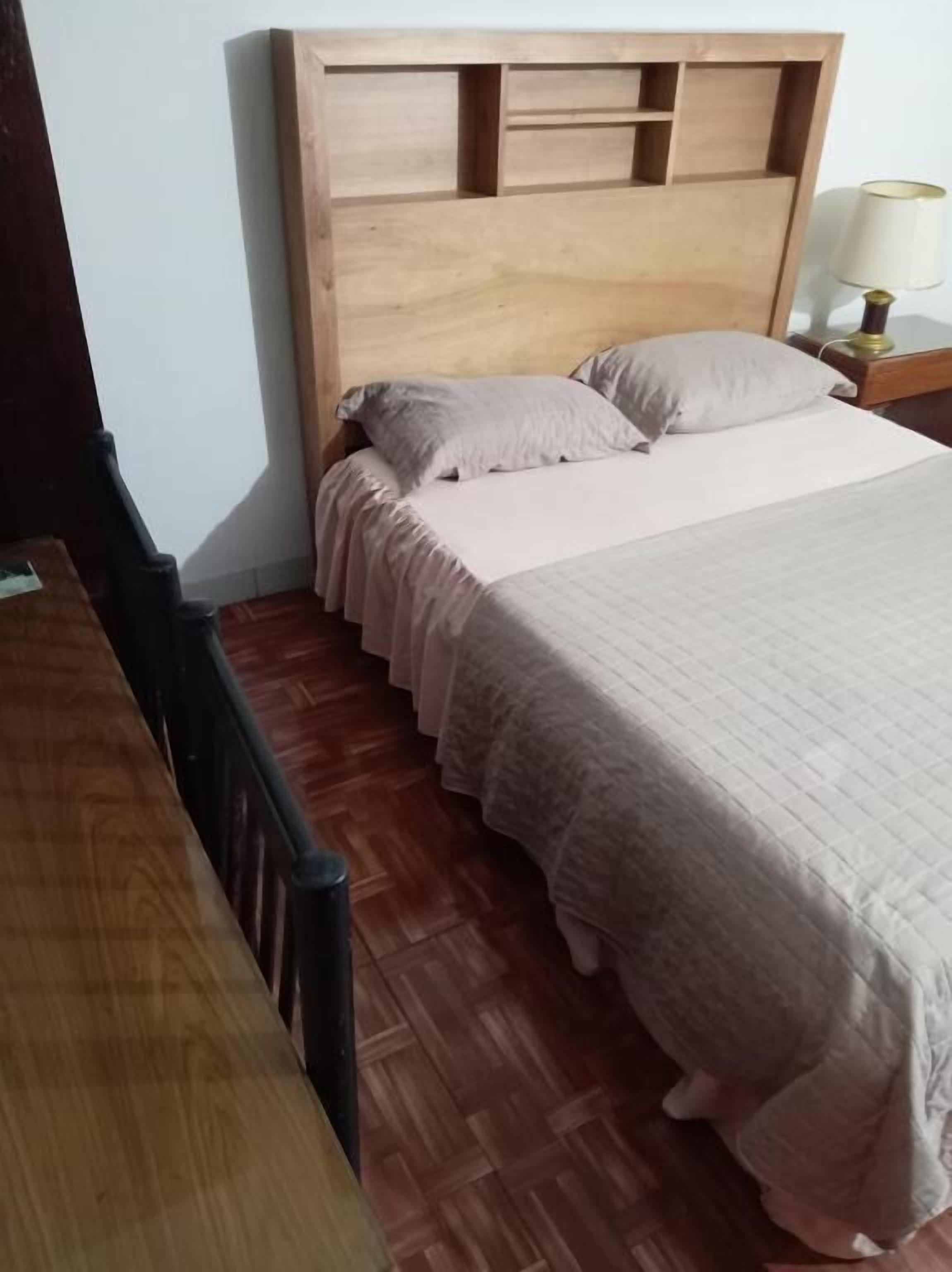 Une chambre à coucher avec une tête de lit en bois, un lit recouvert d’une couvre-lit de couleur claire et une table de chevet avec une lampe.