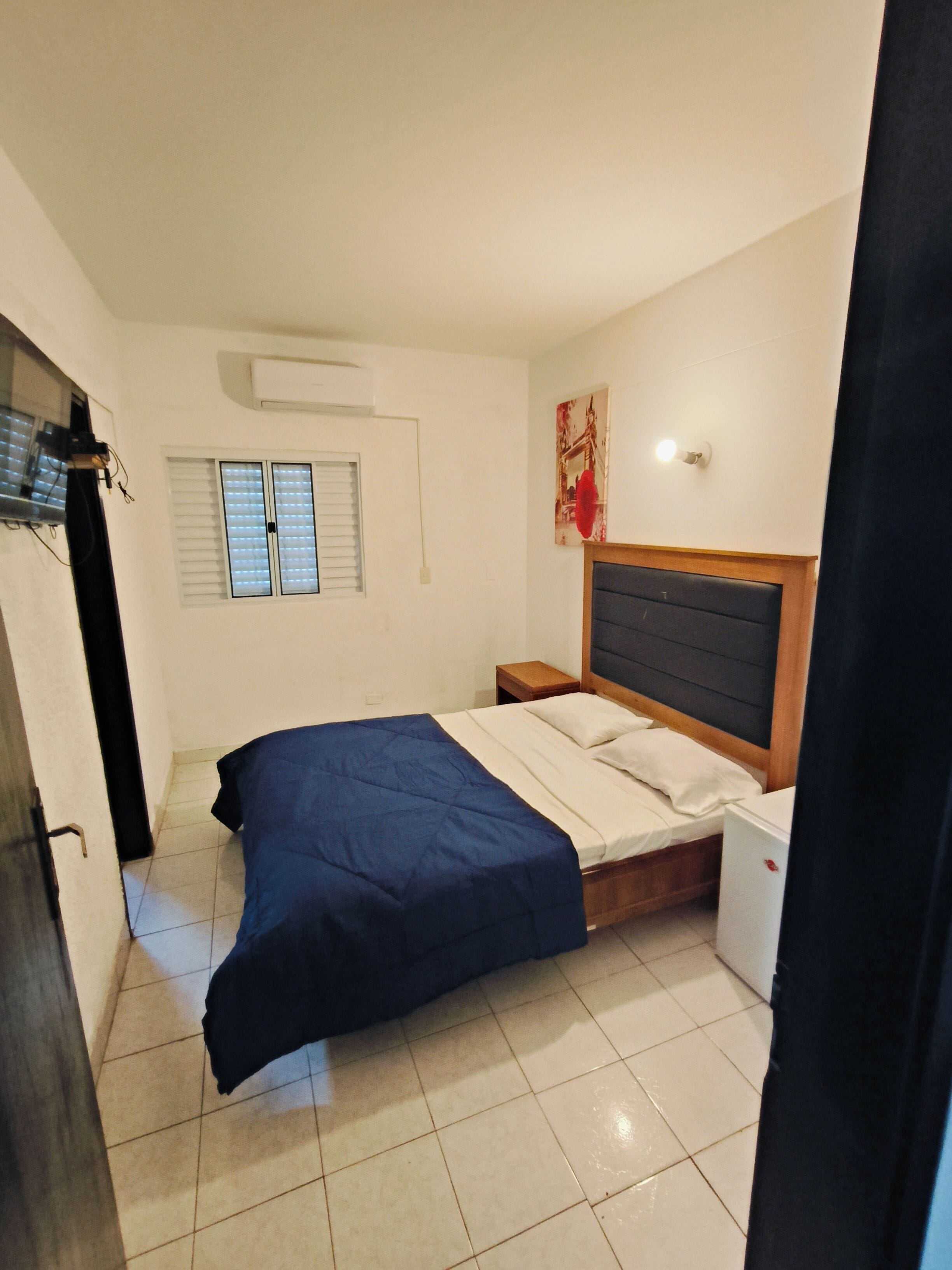 Une chambre à coucher comprenant un lit, une table de chevet, un climatiseur fixé au mur, une fenêtre avec des stores et une porte.