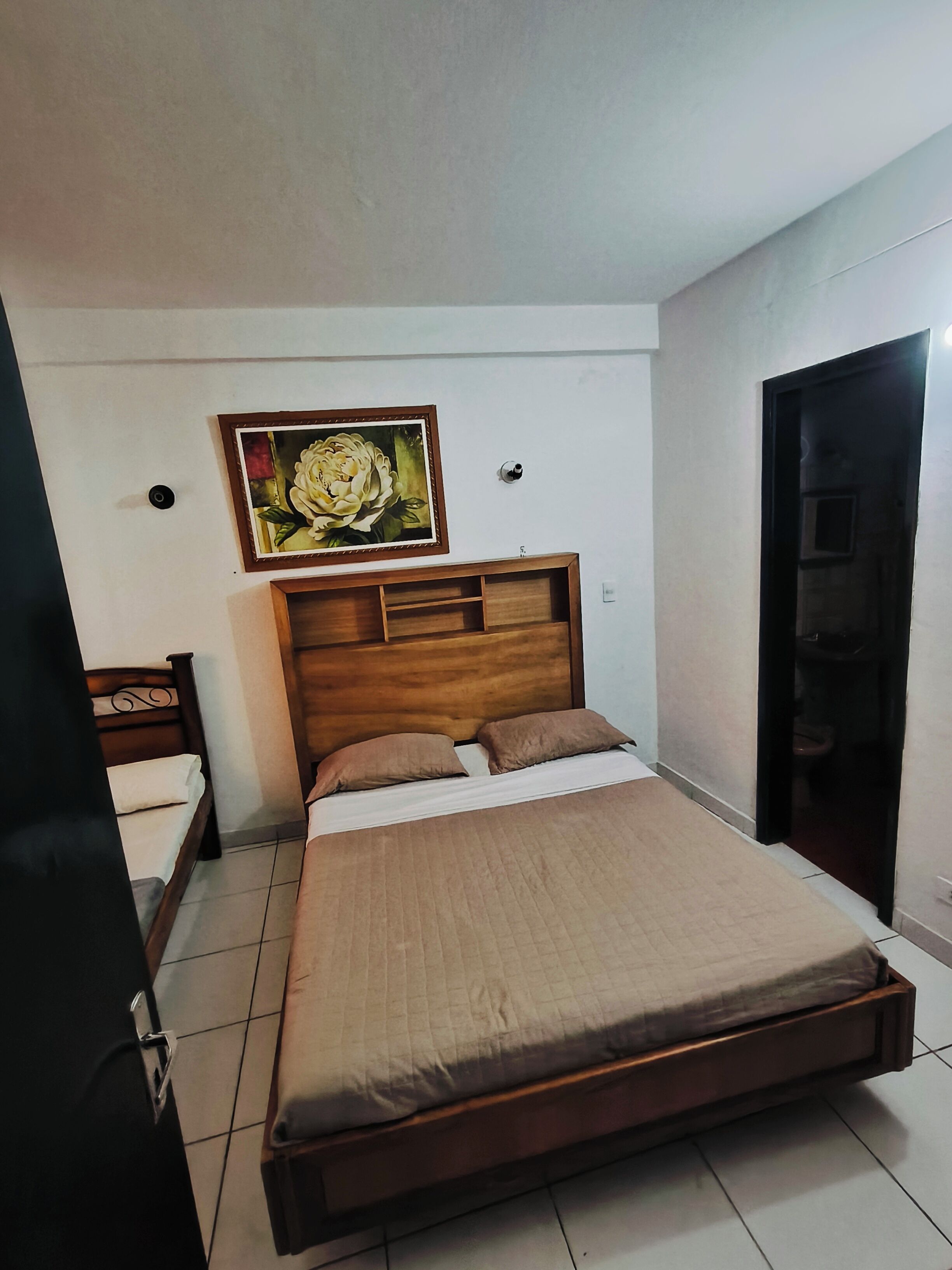 Un dormitorio con una cama de madera, una mesita de noche, un cuadro colgado en la pared, una puerta y un piso con baldosas.